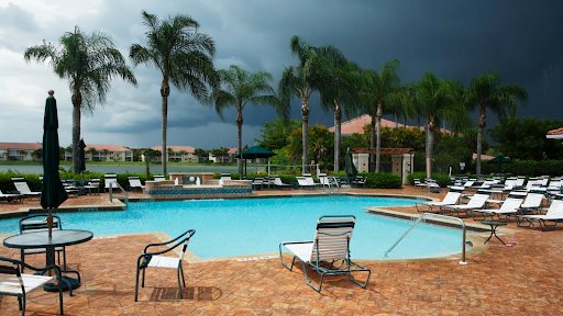 An empty outdoor pool surrounded by chairs and palm trees is shown as dark gray storm clouds approach.